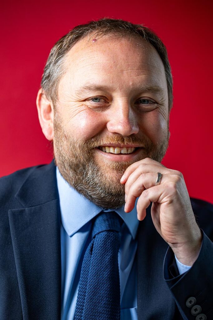Portrait headshot of Labour MP Ian Murray, he is looking into the lens, wearing a navy suit. the background is red. he is smiling and his hand is on his chin. Portrait by Scott Cameron Baxter Aberdeen press and headshot photographer.