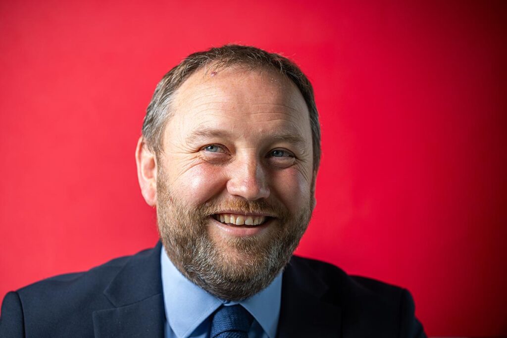 Portrait headshot of Labour MP Ian Murray, he is looking into the lens, wearing a navy suit. the background is red. Portrait by Scott Cameron Baxter Aberdeen press and headshot photographer.